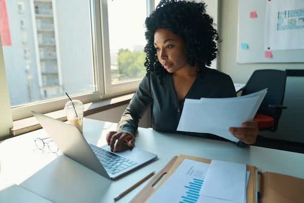 woman at laptop in front of window holding papers